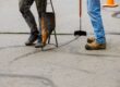 Two construction workers apply sealcoat to asphalt road.