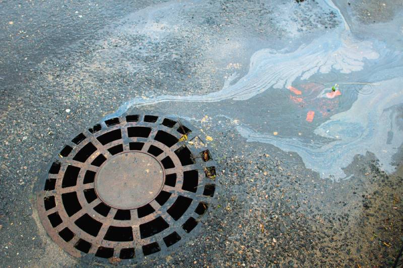 Oil slick on asphalt road drains into a storm drain.